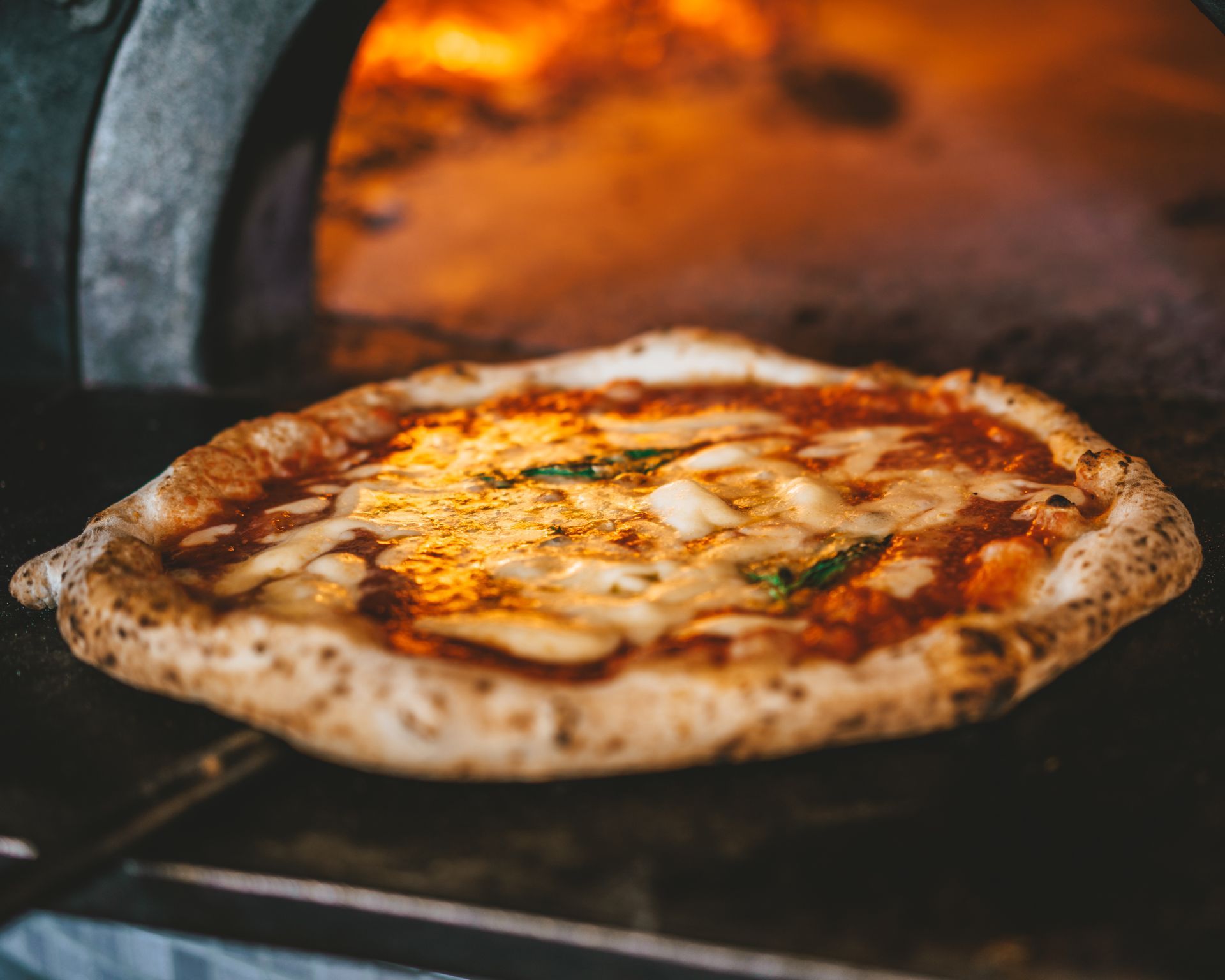 Pizza en train de cuire dans un four à bois ; croûte dorée, fromage fondu, flammes visibles en arrière-plan.
