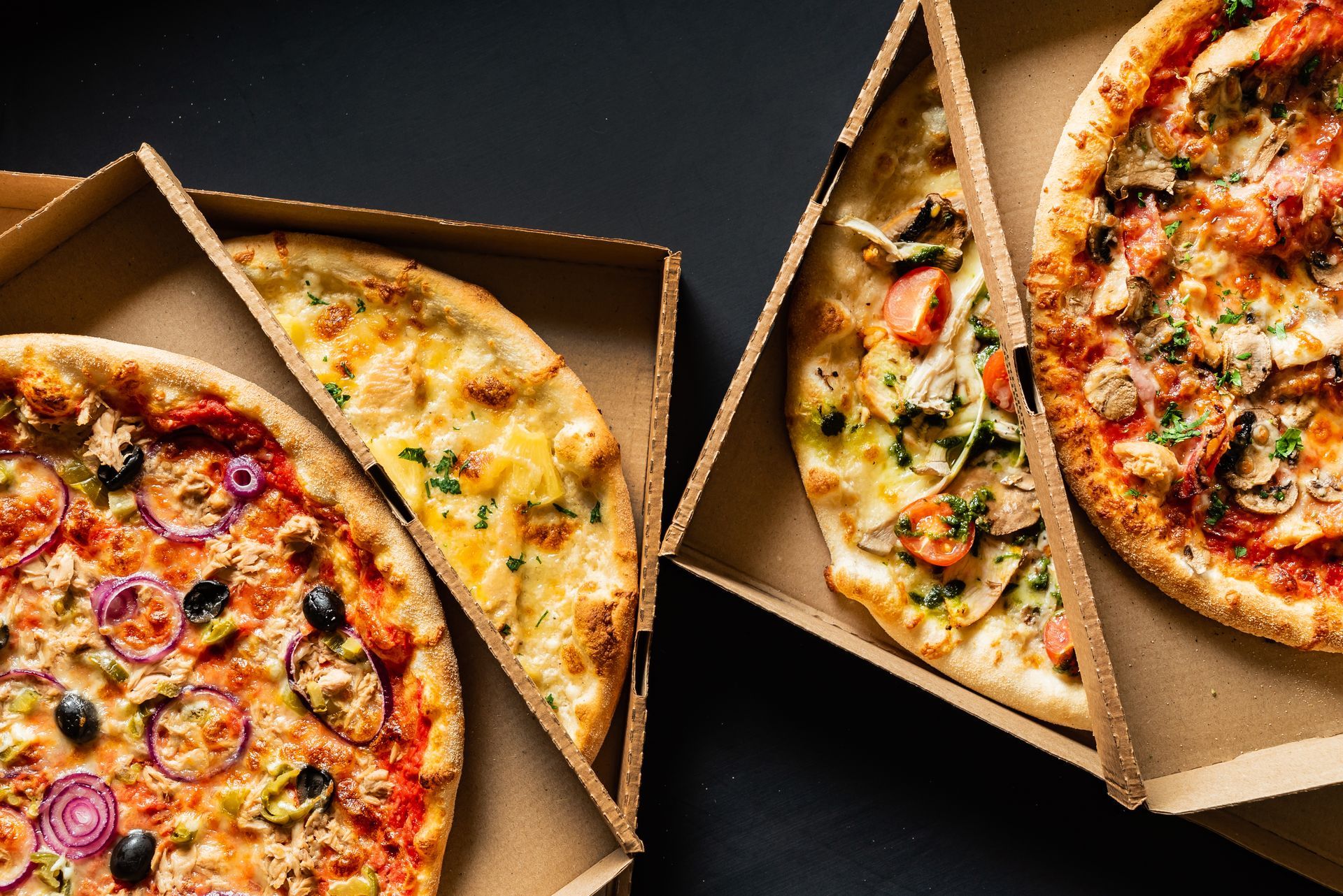 Quatre pizzas dans des boîtes en carton ouvertes, posées sur une surface sombre.