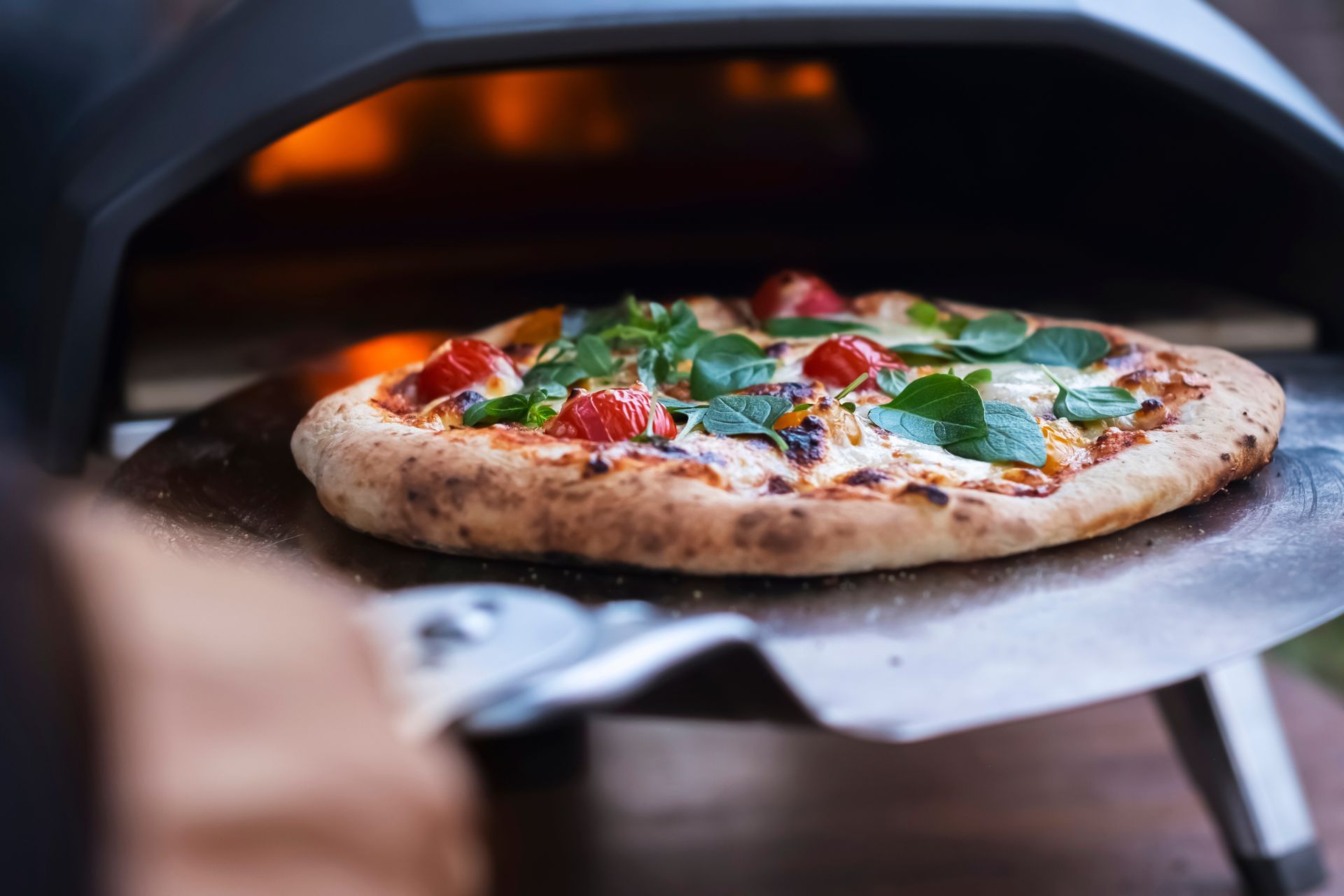Une pizza sur une pelle à pizza, garnie de tomates, de fromage et de verdure, est enfournée dans un four à pizza.