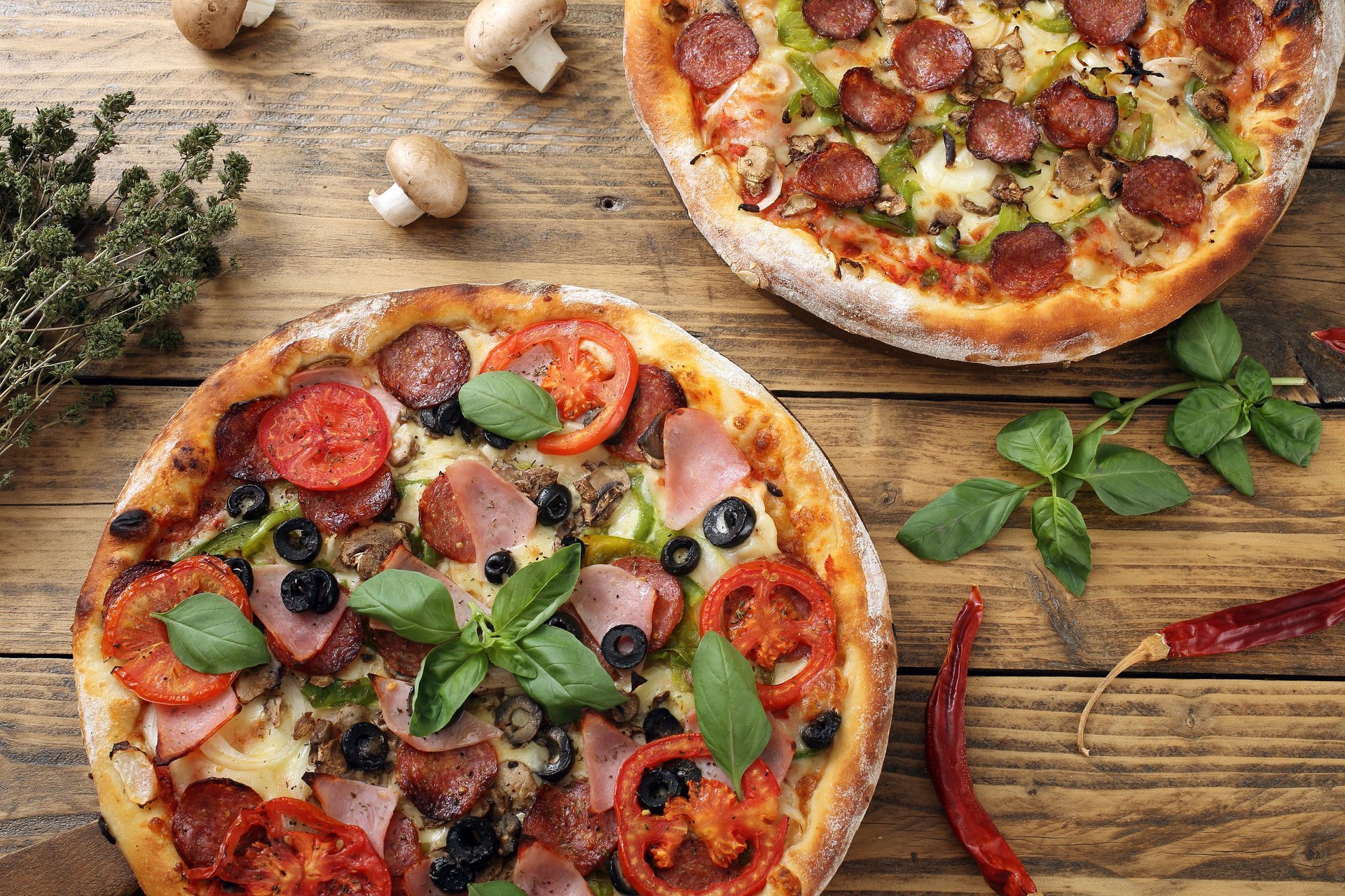Deux pizzas aux garnitures variées sur une table en bois, entourées d'herbes aromatiques et de champignons.