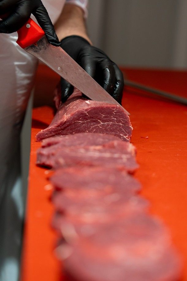 Des mains gantées découpent de la viande crue sur une planche à découper orange avec un couteau