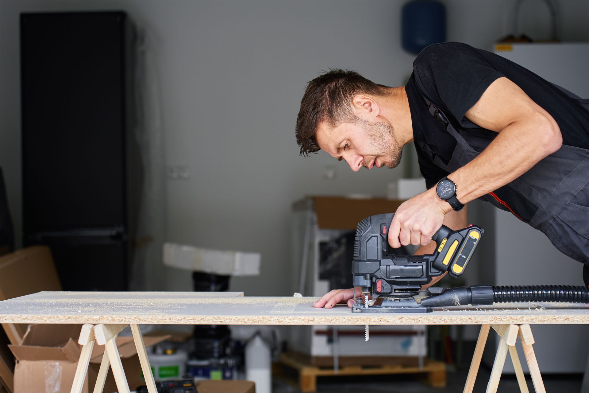 Un homme utilise une scie sauteuse sur une planche en bois posée sur des tréteaux dans un atelier.
