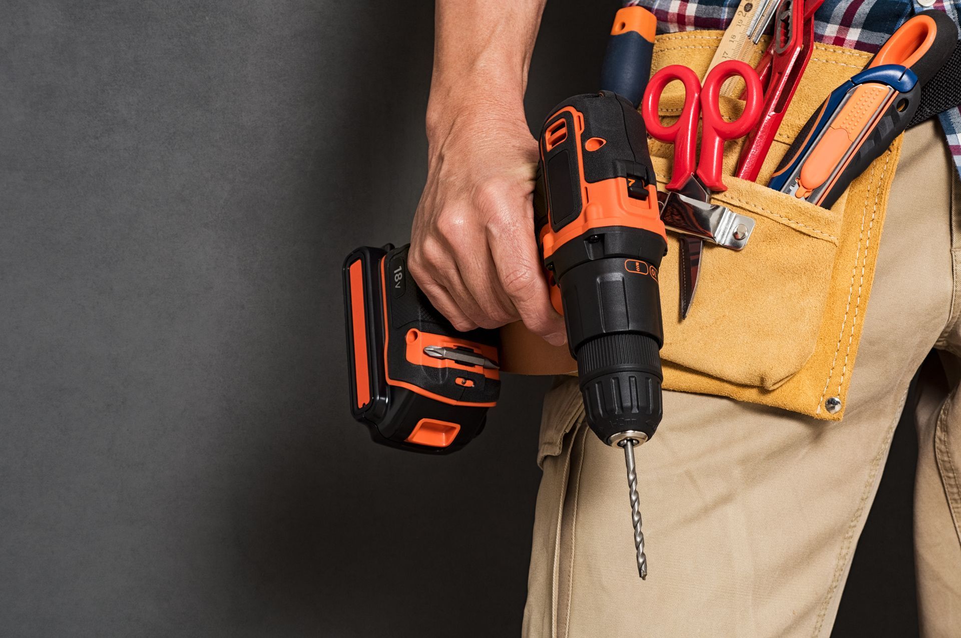 Main tenant une perceuse noir et orange, avec une ceinture porte-outils contenant des ciseaux et des outils.