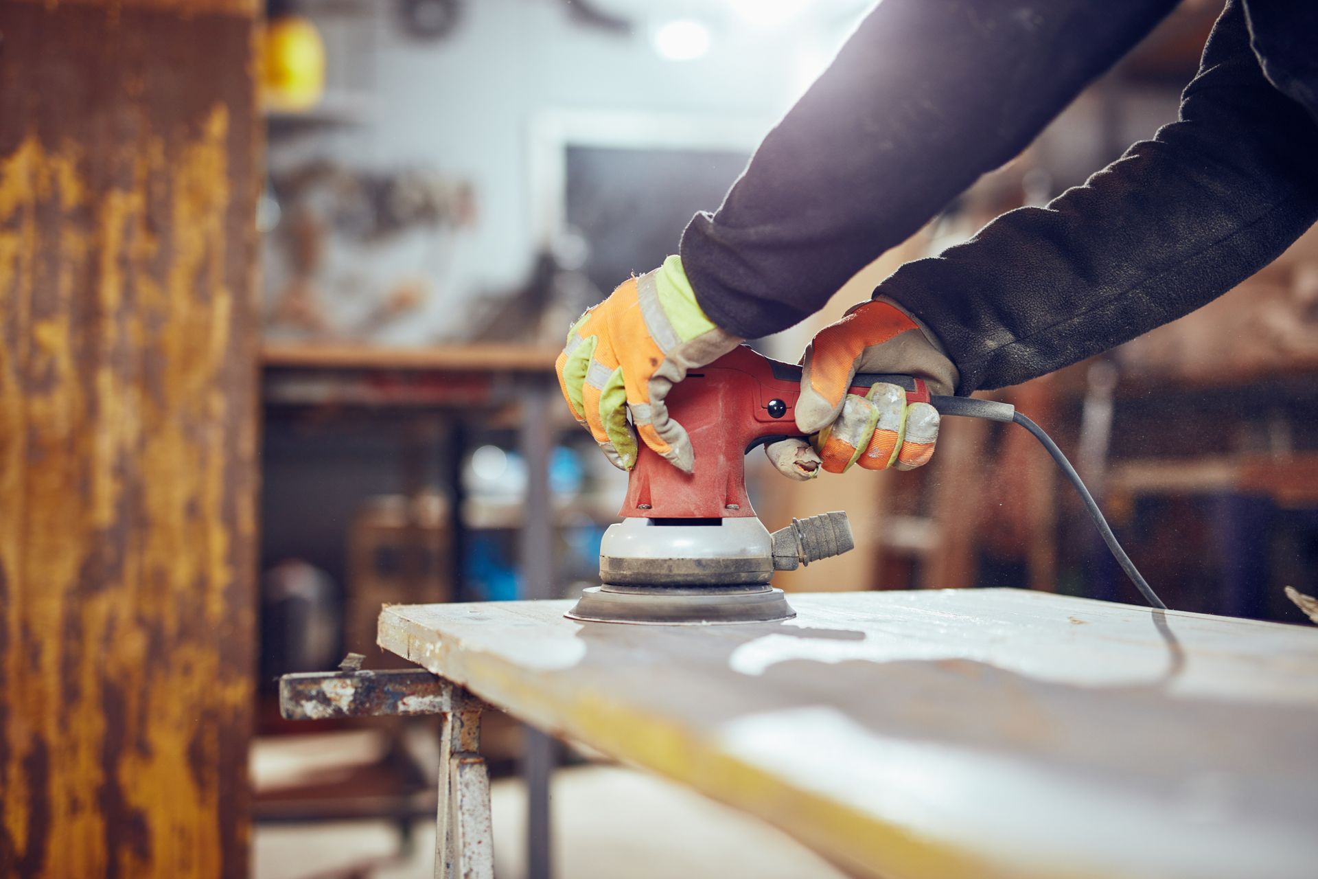 Personne utilisant une ponceuse rouge sur une planche en bois ; portant des gants orange. Atelier.