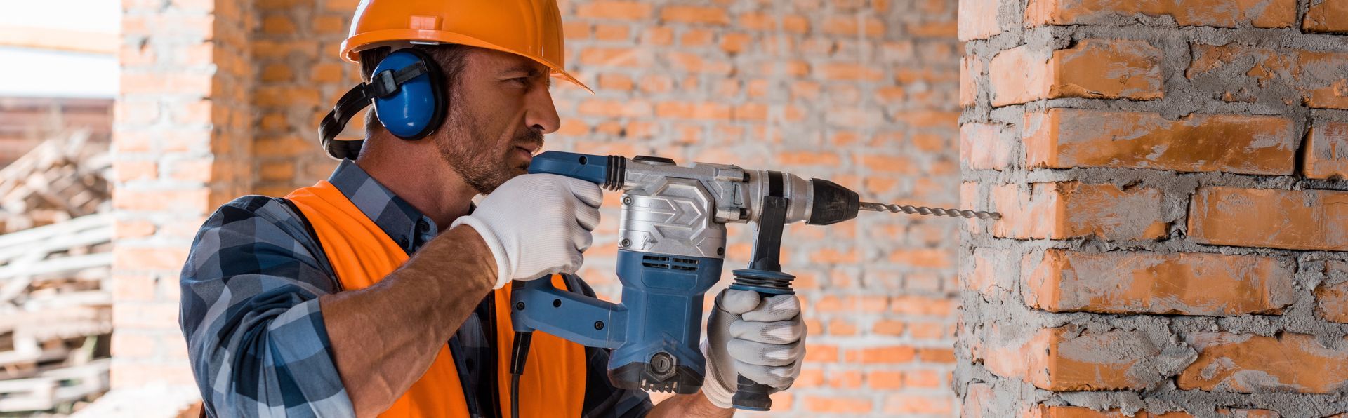 Un ouvrier du bâtiment perce un mur de briques. Il porte des protections auditives, des gants, un casque et un gilet de sécurité orange.
