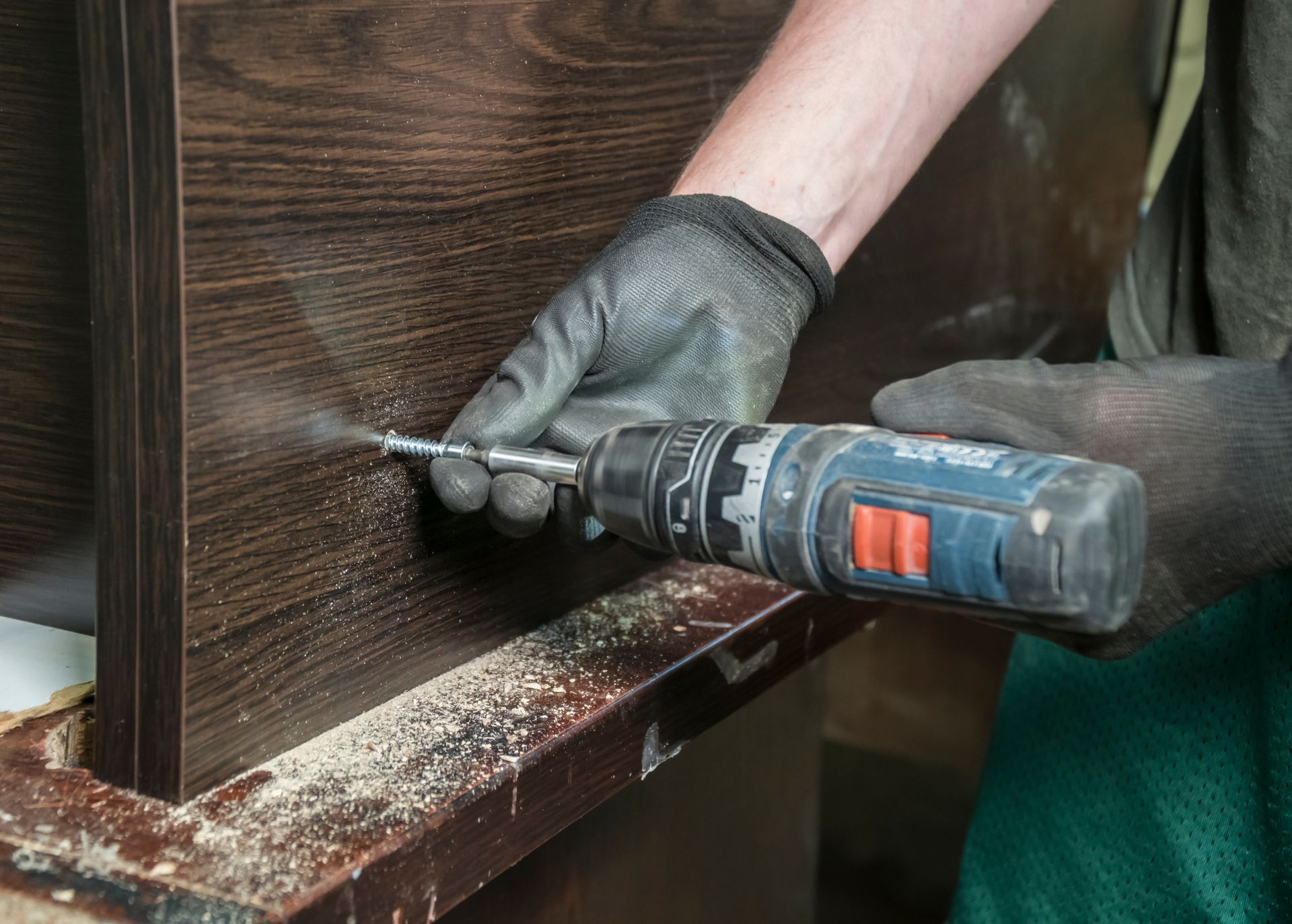 Une personne portant des gants utilise une perceuse électrique sur une armoire en bois ; de la sciure de bois est visible.