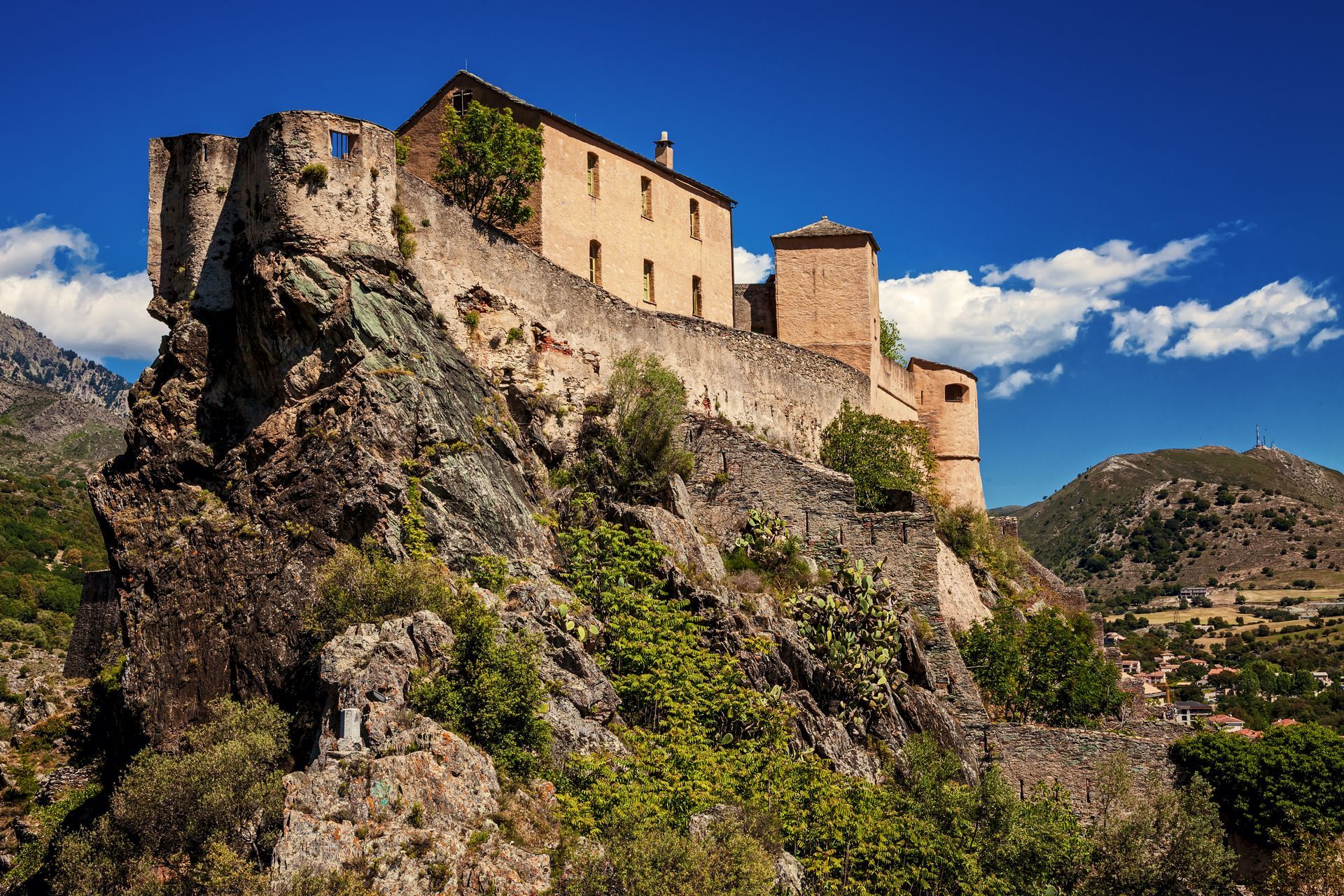 Forteresse en pierre et bâtiment beige au sommet d'une colline rocheuse sous un ciel bleu parsemé de nuages.