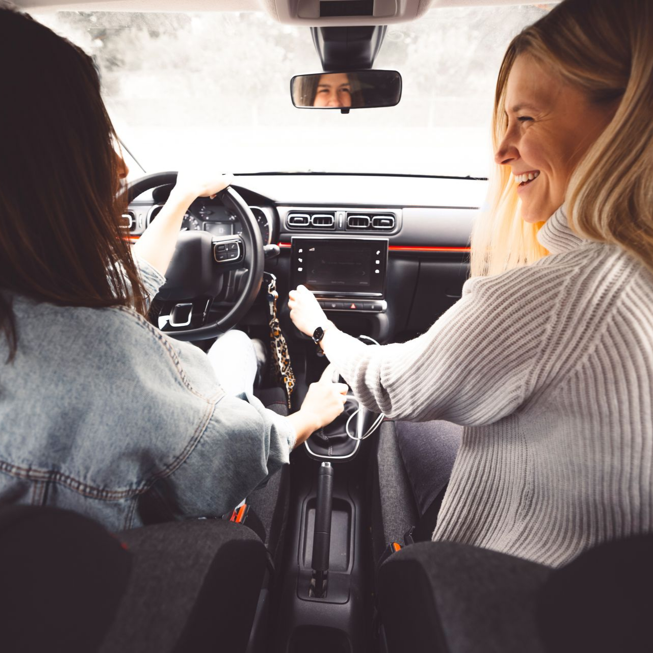 Deux femmes dans une voiture sourient et discutent, l'une au volant et l'autre tourné vers elle depuis le siège passager.