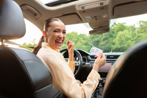 Une conductrice souriante, tenant une carte de permis de conduire assise dans sa voiture, avec une route ensoleillée et des arbres verdoyants à l'extérieur, par la fenêtre.