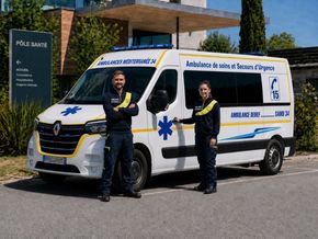 Une ambulance est stationnée devant un hôpital, deux infirmiers en uniforme se tenant à côté.