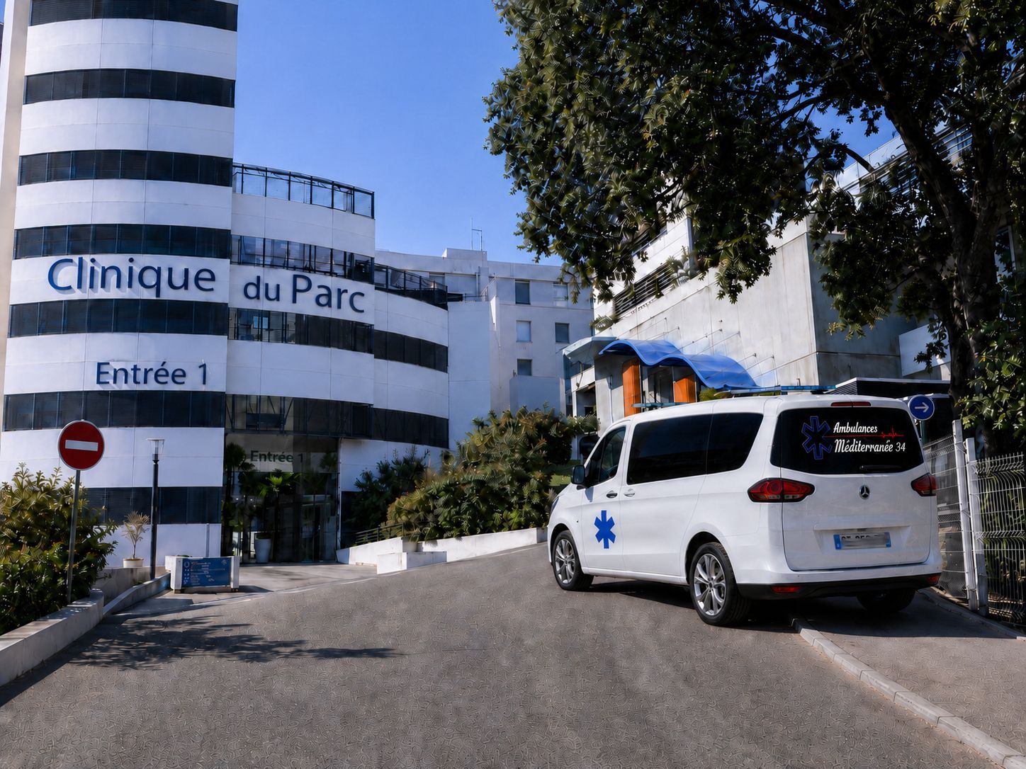 Entrée de la clinique avec une ambulance blanche garée devant un bâtiment blanc sous des arbres.