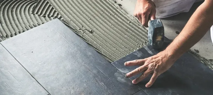 Una persona coloca una baldosa grande y oscura en su sitio sobre una capa de mortero peinado con un mazo de goma.