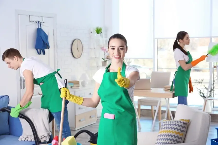 Tres limpiadoras profesionales con delantales verdes y guantes amarillos trabajando en un salón luminoso y moderno.