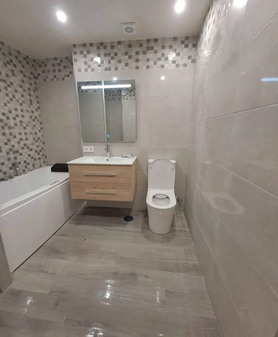 Baño moderno con bañera blanca, lavabo de madera suspendido, espejo e inodoro blanco sobre paredes alicatadas.