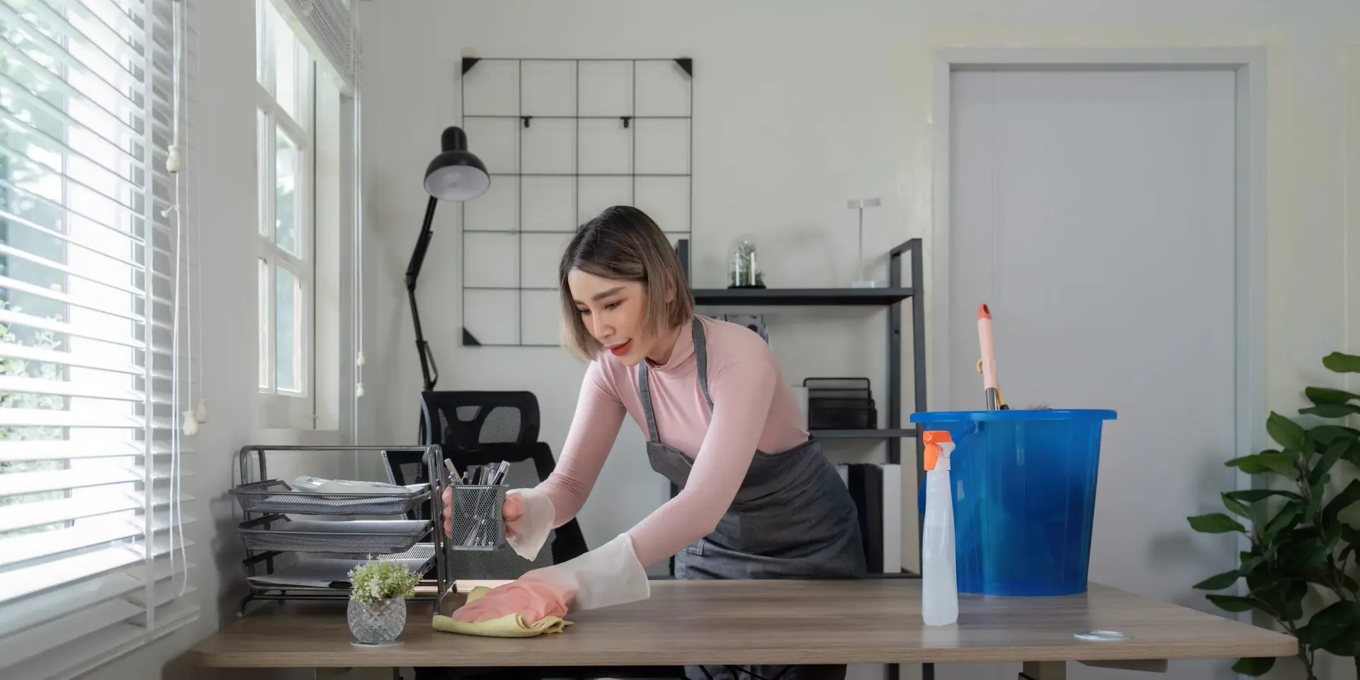 En una oficina luminosa y moderna, una persona con delantal y guantes limpia una mesa de madera con un paño.