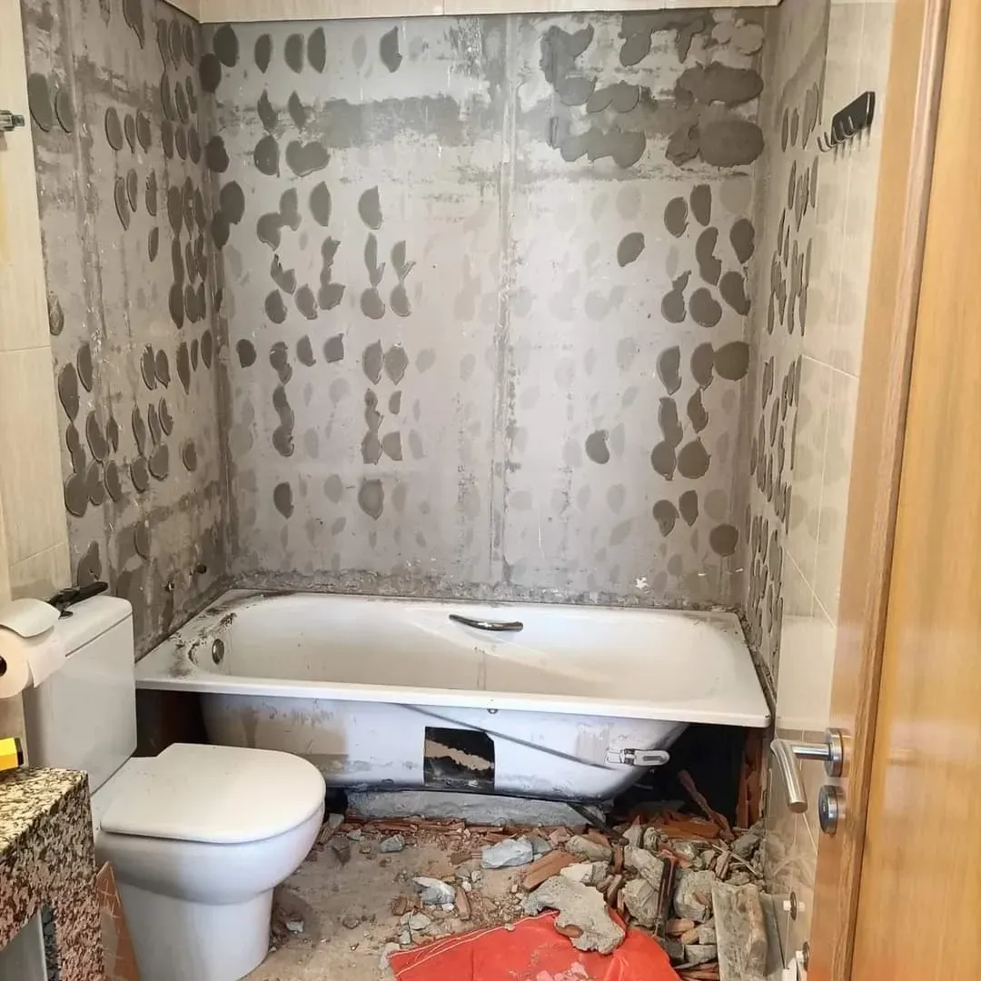 Un baño en proceso de renovación con paredes de hormigón visto, una bañera blanca y escombros en el suelo cerca del inodoro.