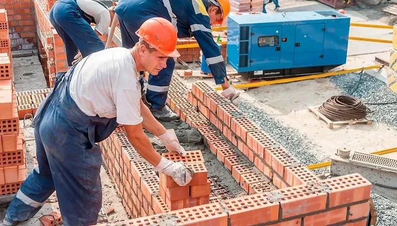 Obreros de la construcción colocan ladrillos rojos para un muro en una obra cerca de un generador azul.