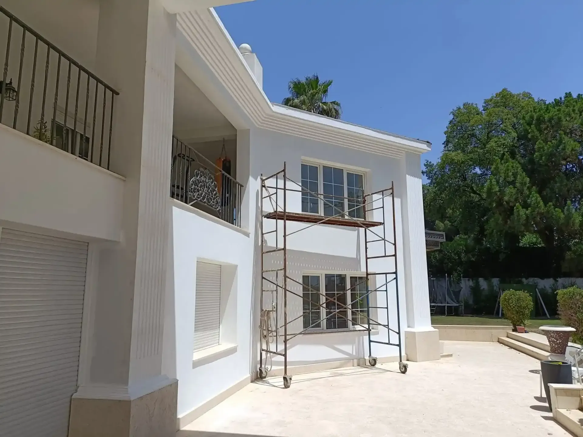 Casa blanca de dos plantas con andamios delante, situada en un patio soleado con árboles.