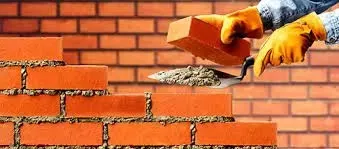 Un albañil con guantes amarillos coloca un ladrillo rojo sobre el mortero con una llana, frente a una pared de ladrillos.