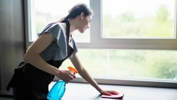 Una persona con delantal limpia una superficie junto a una gran ventana con un pulverizador y un paño.