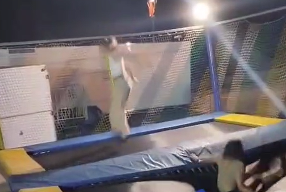 Una persona salta en un trampolín de noche. Lleva ropa clara. Redes de seguridad rodean el trampolín.