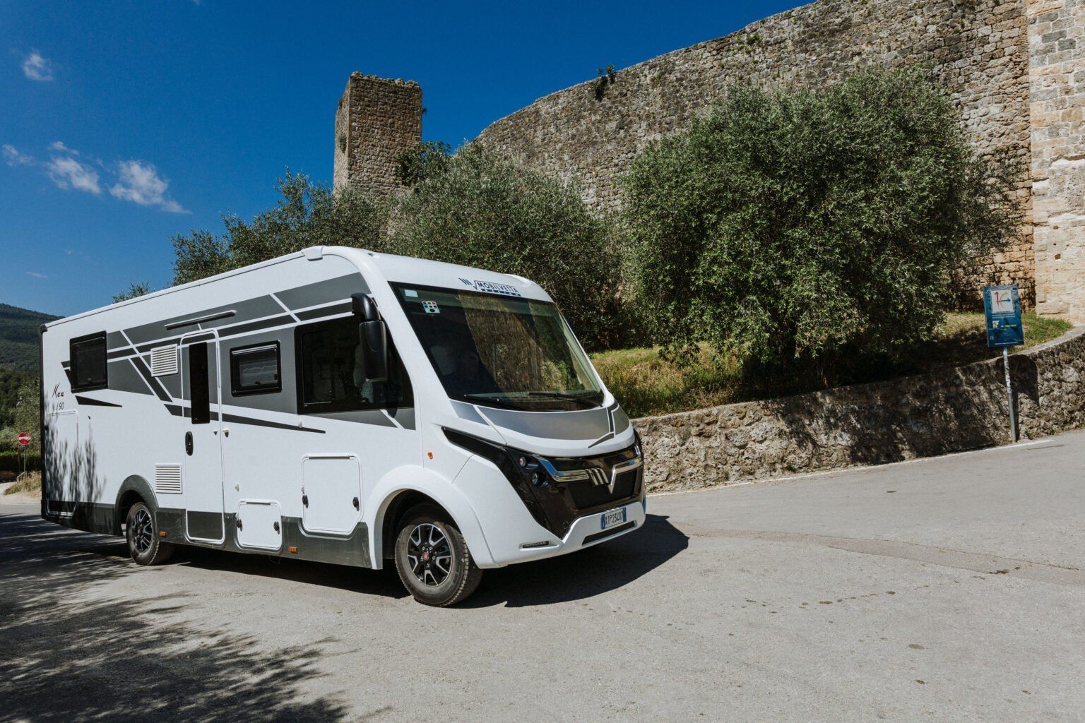 Photo de 3/4 d'un camping-car en train de rouler sur une petite route en cote au pied d'un mur médiéval.