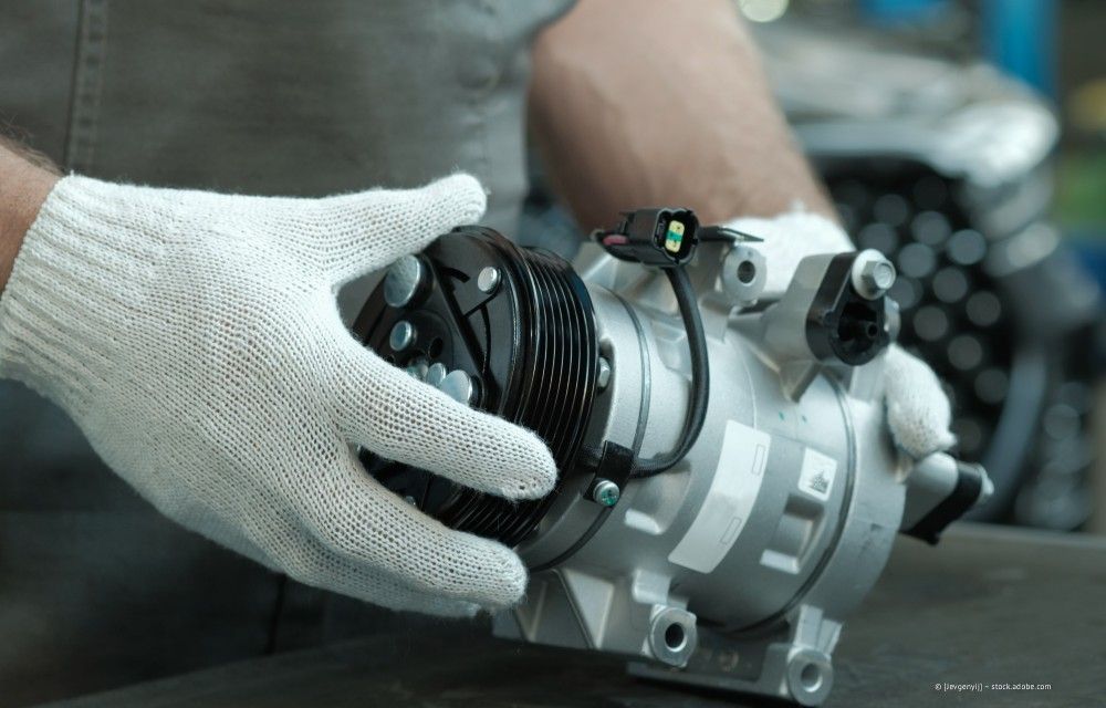 Behandschuhte Hände halten in einer Werkstatt ein silbernes mechanisches Bauteil mit einem schwarzen, fächerartigen Element.