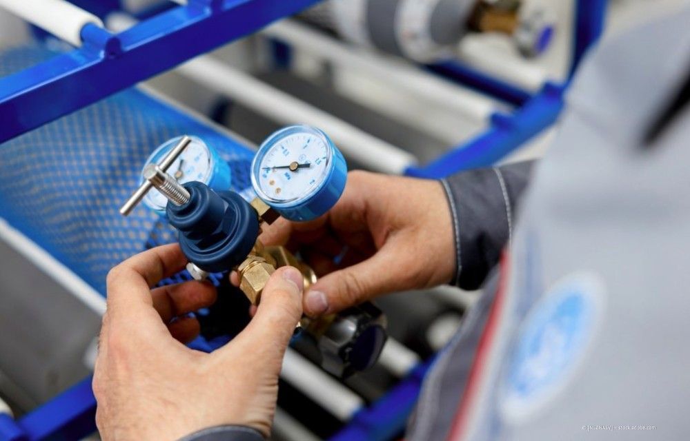 Hände justieren einen blauen Druckregler mit zwei Manometern in einer industriellen Umgebung