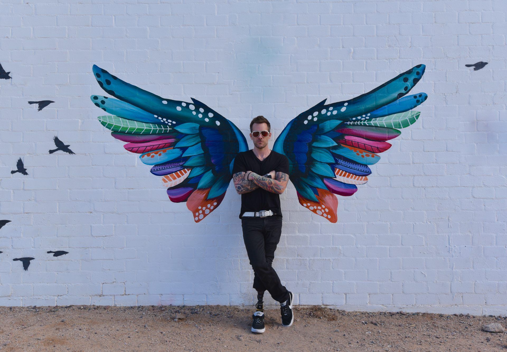 Eine Person steht vor einer weißen Wand aus bunten Vogelflügeln auf dem Rücken, umgeben von fliegenden schwarzen Vögeln