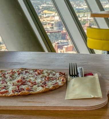 Flammkuchen auf einem Holzbrett mit Besteck, vor einem Fenster mit Blick auf die Stadtlandschaft.