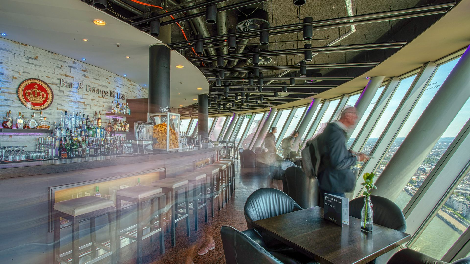 Vogelperspektive auf eine runde Bar mit Panoramafenstern; ein Mann blickt hinaus auf das Stadtbild.