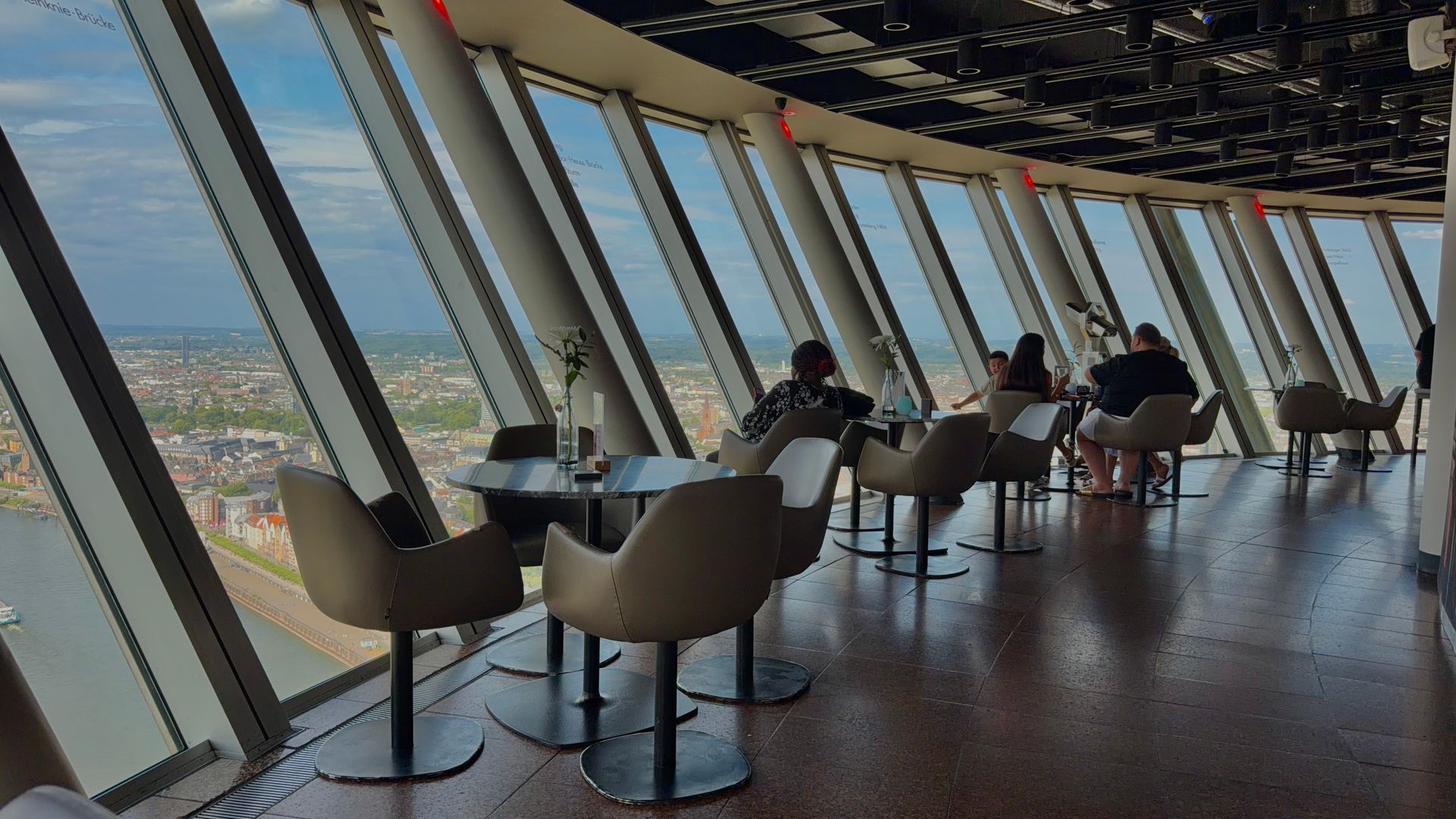 Menschen sitzen in einer modernen Aussichtslounge an großen, schrägen Fenstern mit Blick auf das Stadtbild.