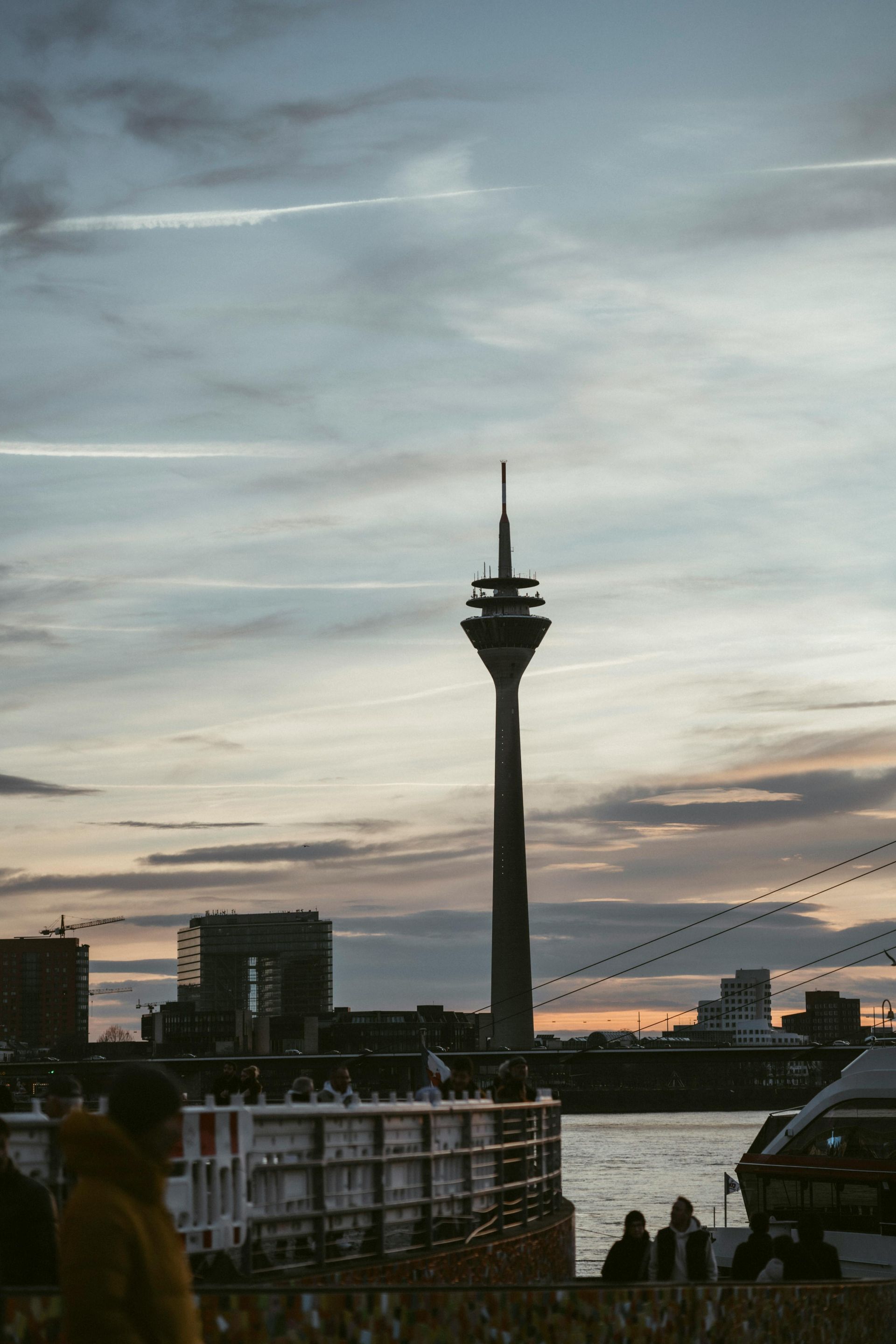 Der Fernsehturm in Düsseldorf, zeichnet sich vor einem bewölkten Sonnenuntergangshimmel ab, im Vordergrund die Uferpromenade.
