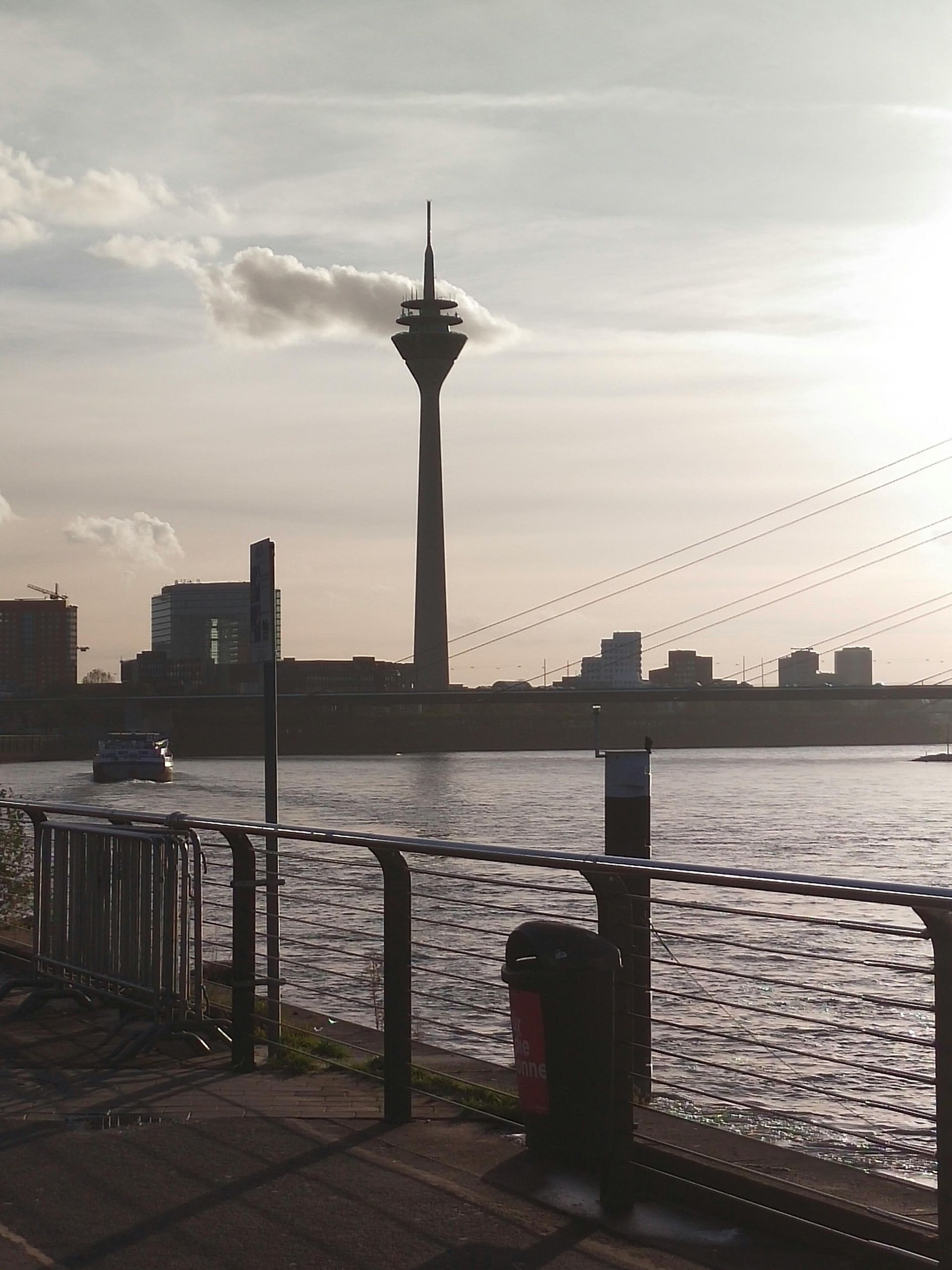 Blick auf den Rhein, mit dem Düsseldorfer Fernsehturm als Silhouette vor bewölktem Himmel. Ein Boot fährt auf dem Wasser.