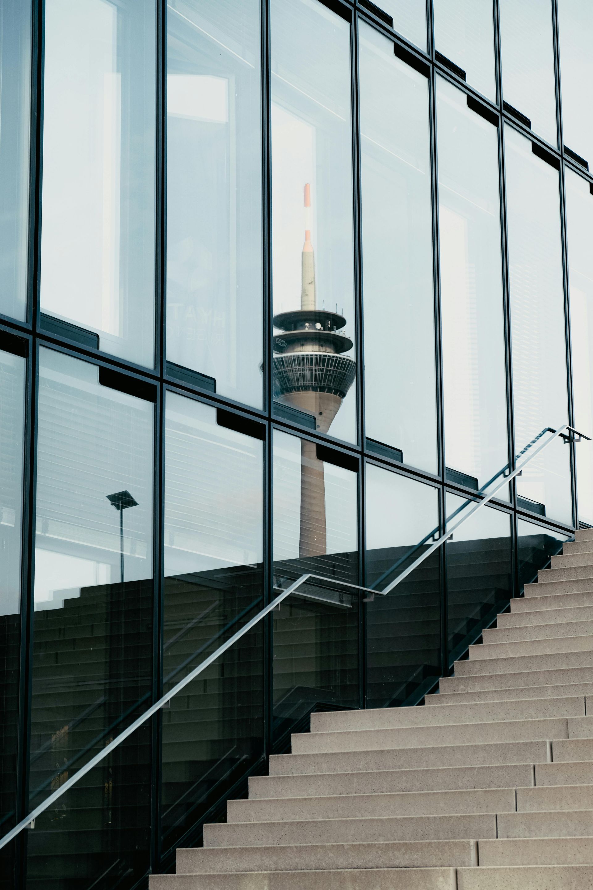 Eine Treppe mit Handlauf führt hinauf zu einem Gebäude mit Glaswänden, in dem sich ein entfernter Fernsehturm spiegelt.