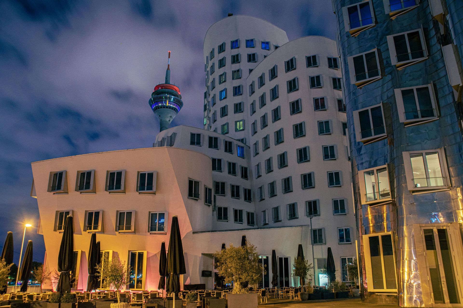 Nachtansicht der Gehry-Gebäude in Düsseldorf, Deutschland, mit beleuchtetem Turm im Hintergrund.