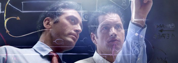 Dos hombres con camisa y corbata analizan ecuaciones en una pizarra transparente en una oficina luminosa.