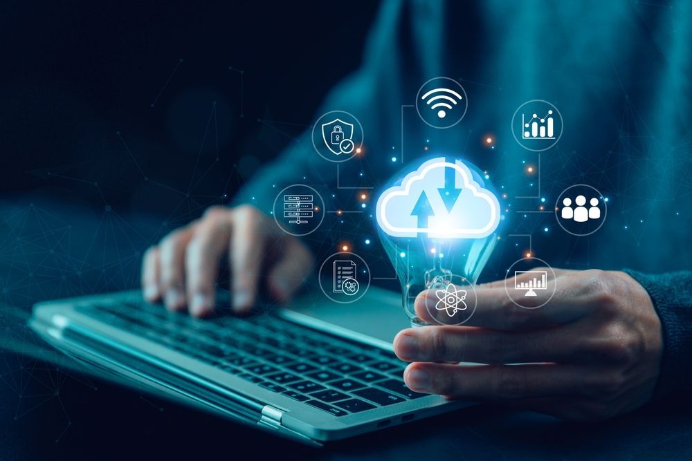Una persona trabajando en un ordenador portátil, sosteniendo el icono de una bombilla brillante con una nube digital flotante y símbolos tecnológicos.