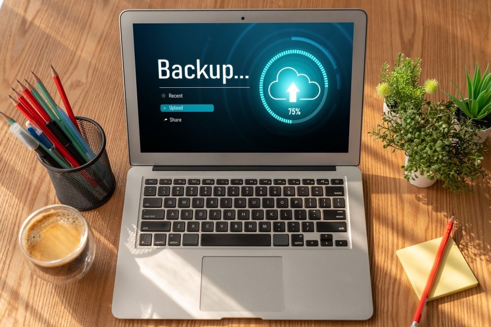 Laptop mostrando el progreso de la copia de seguridad en un escritorio con portalápices, taza de café, planta y cuaderno рядом