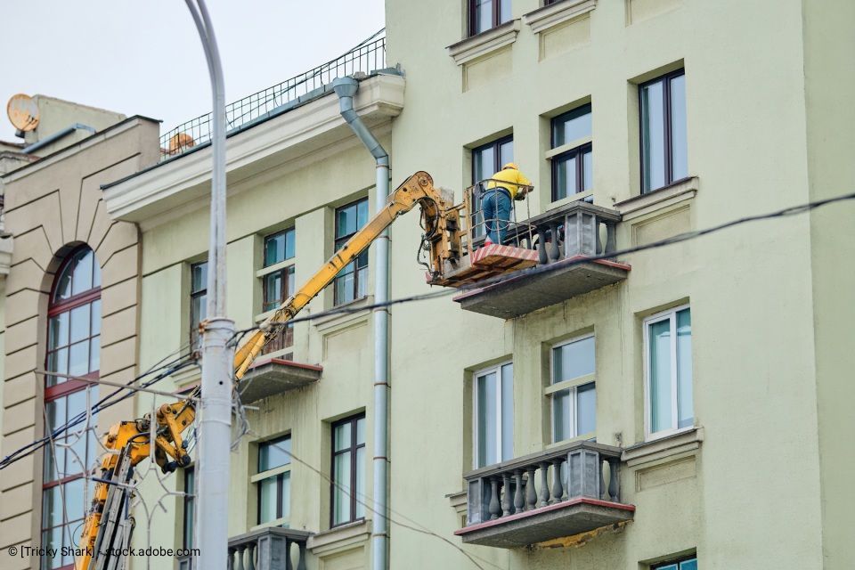 Ein Arbeiter mit Helm steht auf einer Aufzugsplattform nahe der Fassade eines Gebäudes. Der gelbe Aufzugsarm fährt zum Balkon aus.