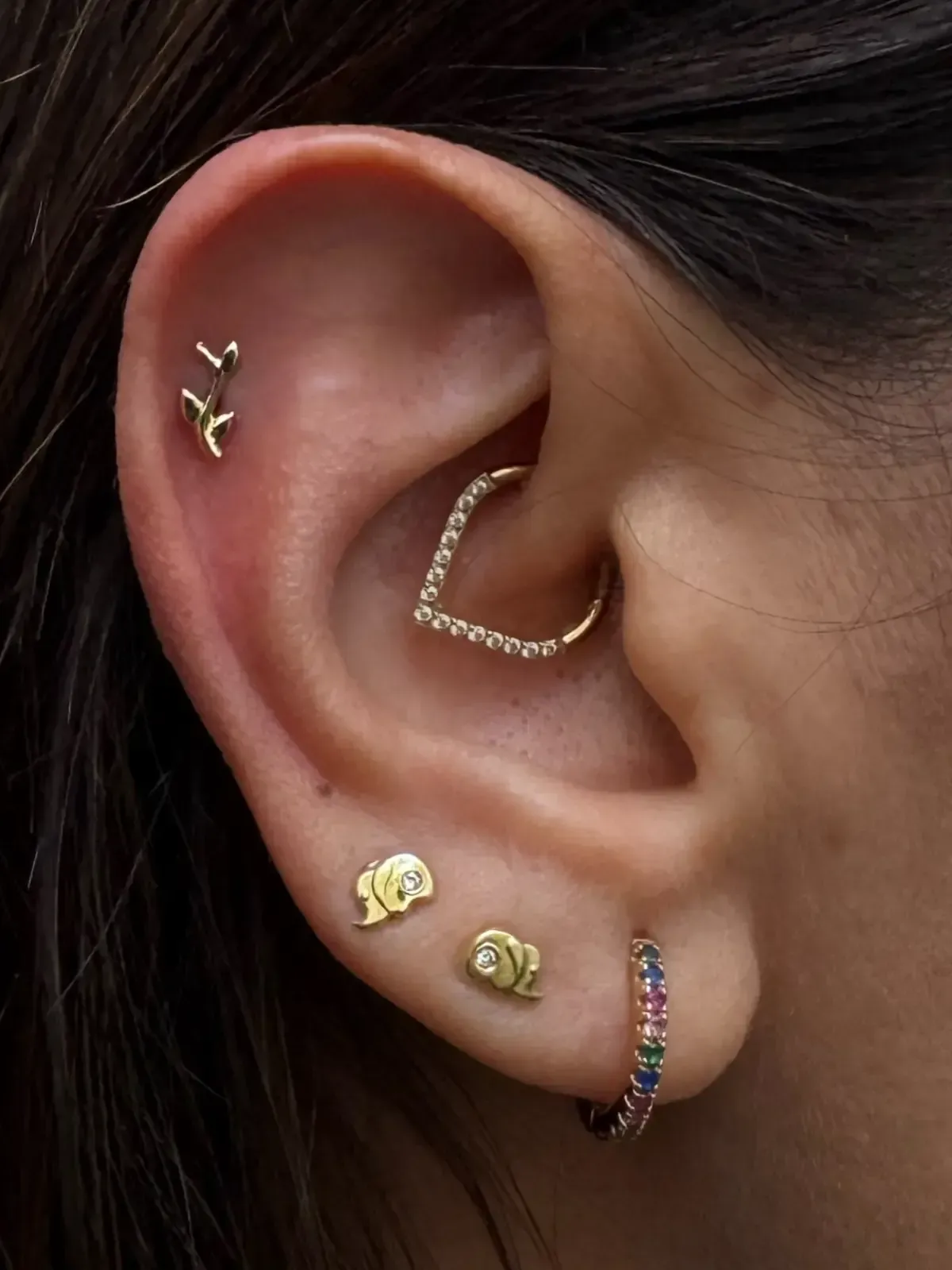 Primer plano de una oreja con múltiples perforaciones de oro, incluido un aro en forma de corazón y pequeños pendientes.
