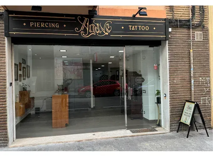 Fachada de tienda de tatuajes y piercings con grandes ventanales, toldo oscuro y cartel en forma de A.