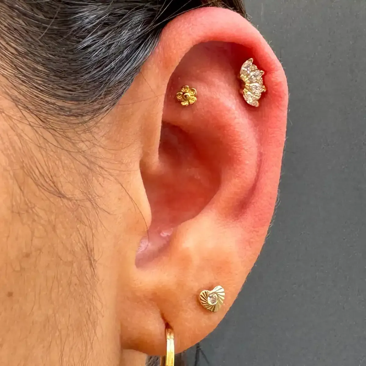 Primer plano de una oreja con tres pendientes de oro: una flor, un racimo y un corazón.