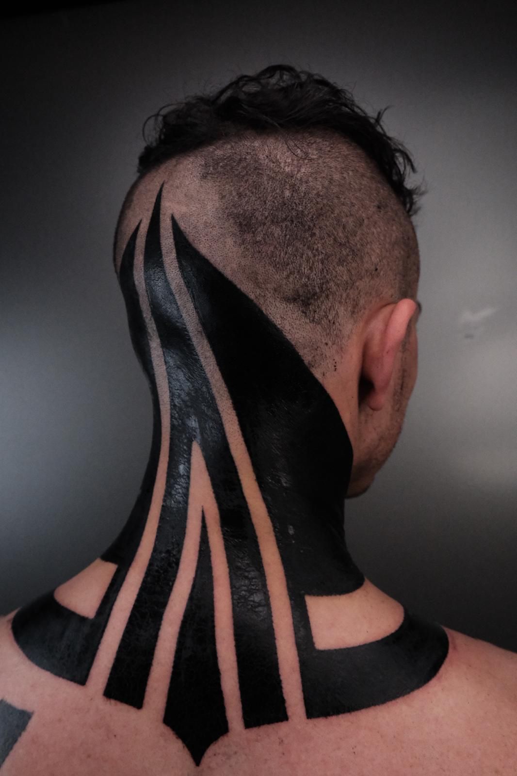 Nuca y cabeza con tatuajes geométricos negros. Cabello corto, laterales rapados y fondo oscuro.