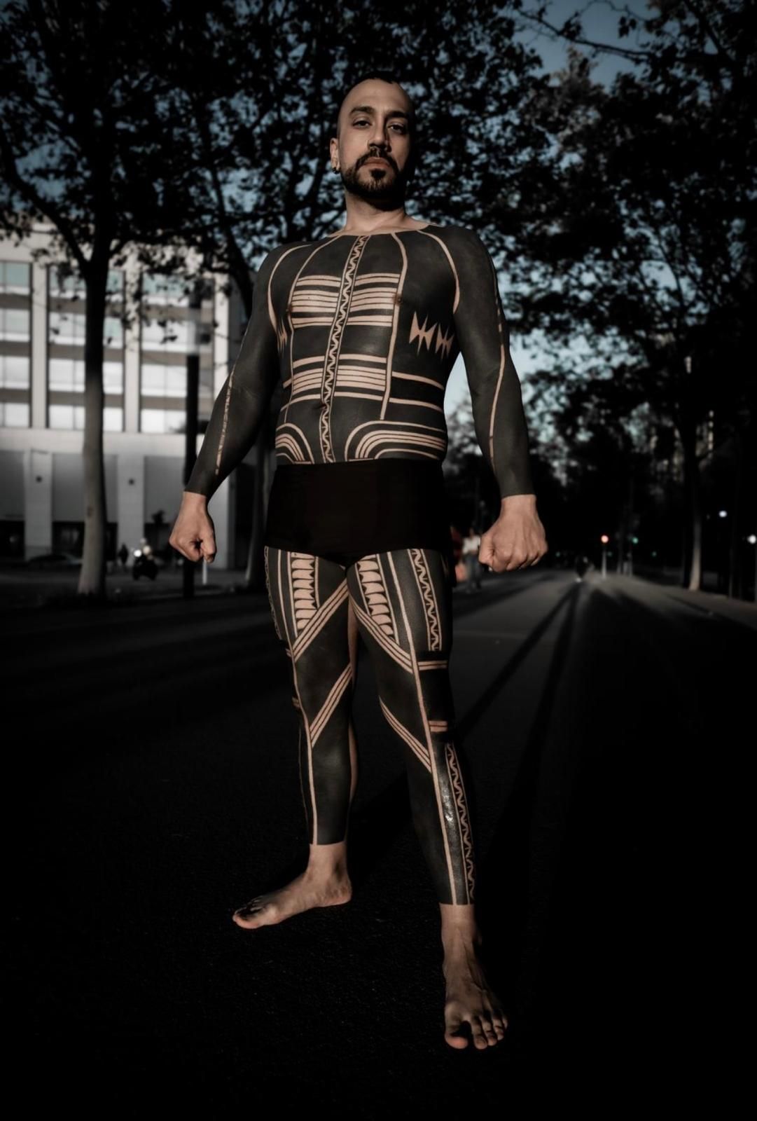 Un hombre con tatuajes negros en todo el cuerpo está de pie en un sendero oscuro. Descalzo, con pantalones cortos negros.