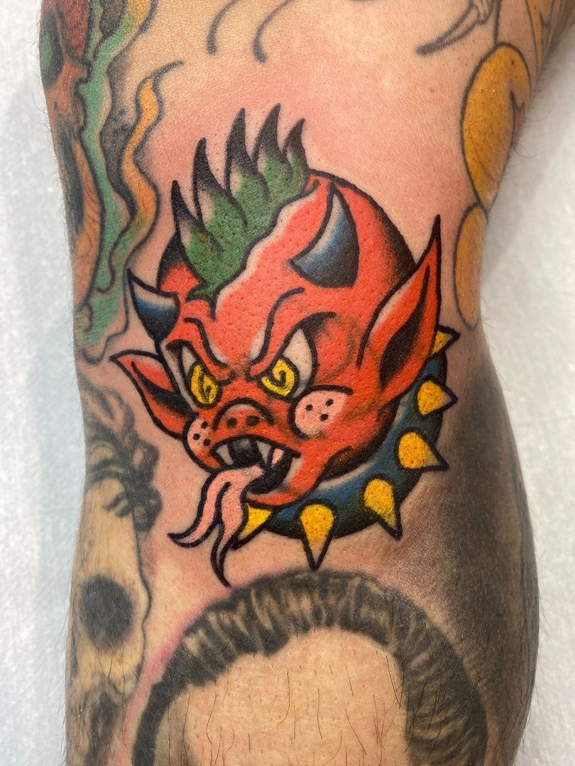 Tatuaje de una cabeza de diablo rojo con cuernos, lengua afuera y collar con púas. Colocado en una pierna.
