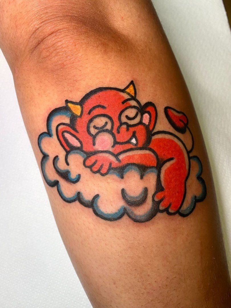 Diablo rojo de dibujos animados durmiendo en una nube azul, tatuado en el brazo de una persona.