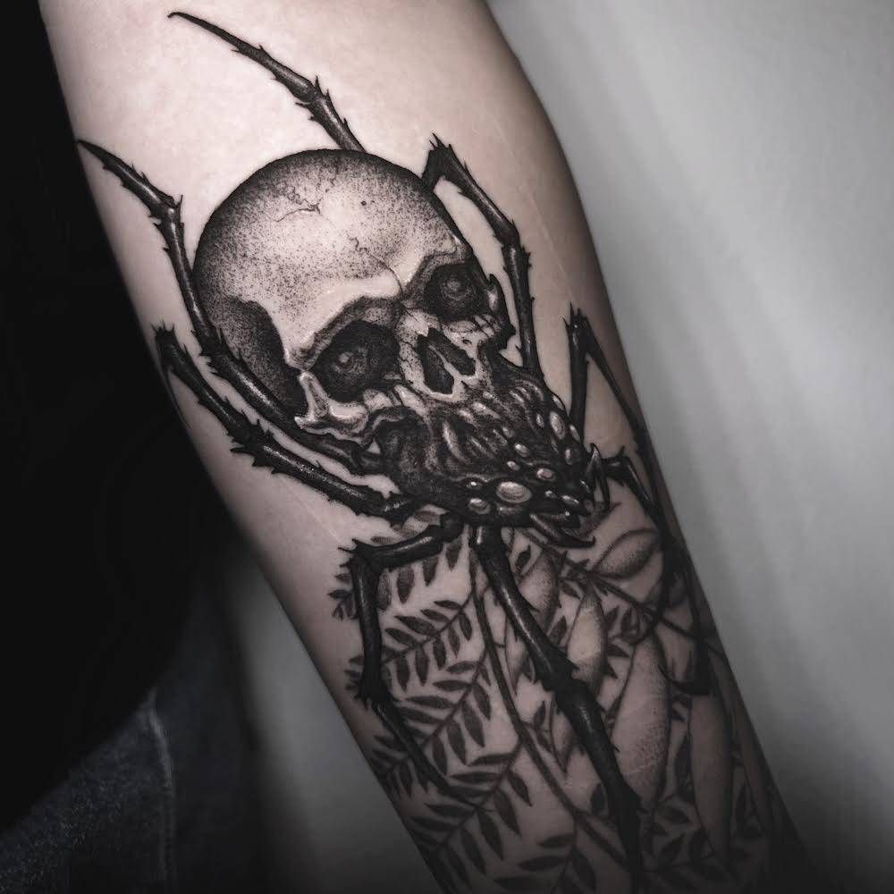 Tatuaje de calavera y araña en el brazo, tinta negra. Cabeza de calavera con patas de araña, telaraña y follaje.