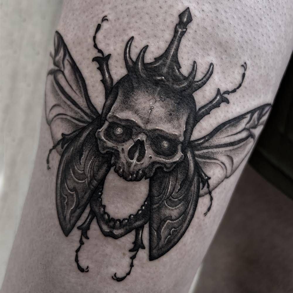 Tatuaje de un escarabajo calavera con mandíbulas abiertas, alas y cuernos en sombreado negro y gris.