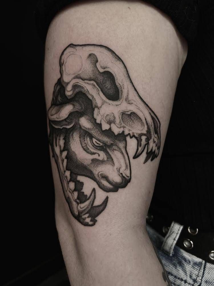 Tatuaje de una calavera de perro sobre una cabeza de carnero, en negro y gris, en la parte superior del brazo de una persona.